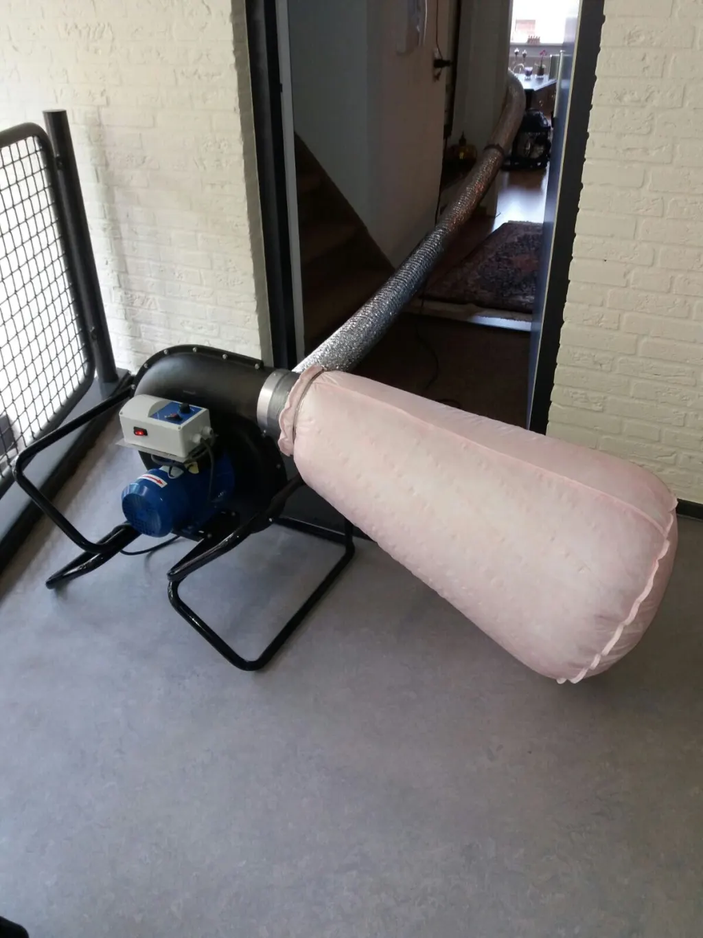 Mobiele stofafzuiger met blauwe motor, zwart slakkenhuis en roze opvangzak op een overloop; een flexibele aluminium slang loopt de woning in